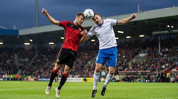 Dos jugadores de fútbol, uno con camiseta roja y otro con camiseta blanca, saltan alto para rematar un balón con la cabeza en un campo verde.