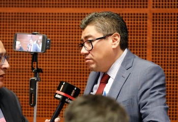 Primer plano de Saúl Monreal Ávila, un hombre con traje azul, corbata roja y gafas, hablando a un micrófono; un smartphone graba en segundo plano