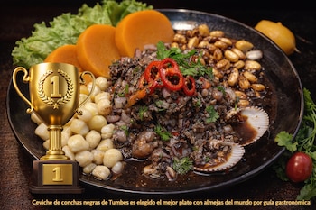 Un plato oscuro contiene ceviche de conchas negras, maíz blanco, camote, lechuga, cilantro y ají rojo. Junto al plato hay una copa trofeo dorada con el número uno.