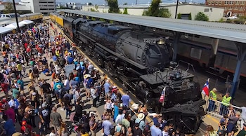 Una locomotora de vapor negra con la inscripción X4014 y una pequeña bandera estadounidense avanza sobre vías en un entorno de árboles verdes