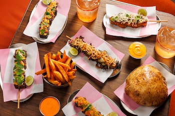 Mesa de madera con brochetas sobre papel rosa, papas fritas, pan horneado, salsas y dos vasos con bebidas con hielo