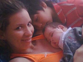 Una mujer sonriente mira a la cámara con un bebé durmiendo sobre su pecho y la cabeza de otra persona visible detrás, todos abrazados