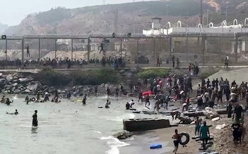 Multitud de personas en playa y mar; algunos individuos escalan una valla fronteriza de metal con concertinas en la parte superior