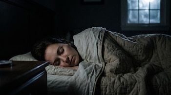 Primer plano de una mujer durmiendo plácidamente de lado en una cama, tapada con sábanas, en una habitación oscura con una ventana al fondo.