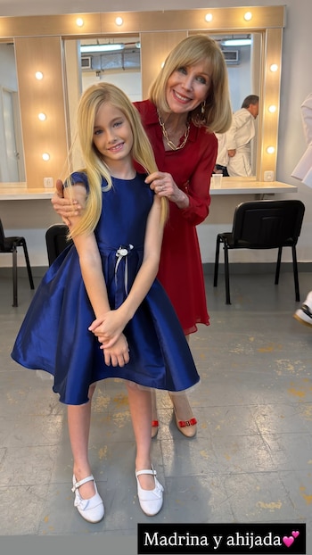 Una niña con vestido azul y zapatos blancos posa de pie junto a una mujer con vestido rojo en un camerino con espejos iluminados