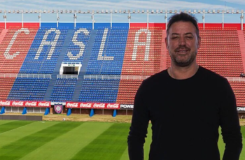 Un hombre de cabello oscuro y barba, vestido con una camiseta negra, posa sonriendo frente a las gradas de un estadio con asientos rojos y azules que forman 'CASLA'