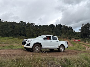 Una Toyota Hilux blanca circula por un camino de tierra y barro. Un conductor es visible. Hay árboles y un cielo nublado de fondo