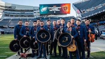 Once músicos vestidos con traje de mariachi azul sostienen sus sombreros e instrumentos en una cancha con una pantalla gigante al fondo.