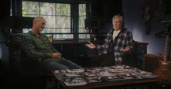 Paul McCartney gesticula mientras conversa con un hombre en un estudio. Una mesa con fotos en blanco y negro los separa, con equipo de audio y guitarras visibles