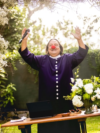 Hombre sonriente con nariz de payaso roja y sotana morada, con un micrófono en mano, de pie detrás de una mesa con laptop, flores y gafas