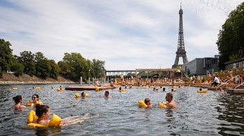 Muchas personas nadan o flotan con flotadores amarillos en el agua del río cerca de plataformas de madera. Se ve la Torre Eiffel y árboles en el fondo.