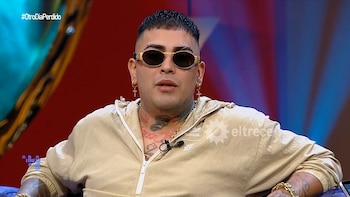 Primer plano de X, un hombre con corte de pelo rapado, gafas de sol ovaladas, piercings y tatuajes en el cuello y brazos, vistiendo una chaqueta beige con cierre