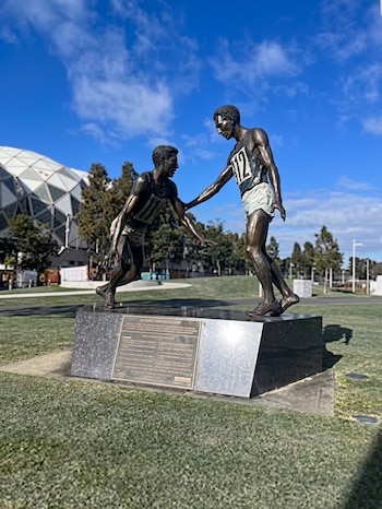 Escultura de bronce sobre pedestal de piedra que representa a dos atletas corriendo en un parque con un estadio al fondo