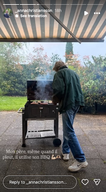 Un hombre de espaldas, con chaqueta verde y jeans, cocina en una parrilla con humo en un patio exterior. Se ve un toldo a rayas y vegetación al fondo