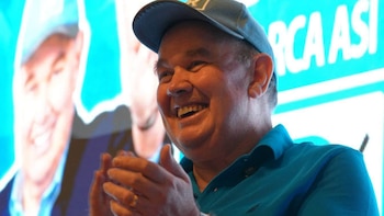 Hombre con gorra y polo azul sonríe y aplaude, frente a una pantalla con texto blanco "RCA ASI" y la imagen borrosa de otro hombre