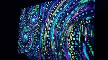 Lienzo de arte abstracto con espirales, puntos y trazos de pintura fluorescente verde, azul y morada sobre fondo negro.