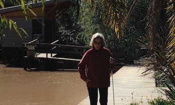 Una mujer de cabello gris con lentes de sol, chaqueta roja, pantalón negro y bastón blanco, camina sobre un deck de madera. Se observa un río, una casa de madera sobre pilotes y vegetación densa