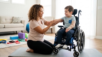 Una mujer arrodillada ayuda a un niño sentado en silla de ruedas con un ejercicio de brazo. El niño tiene una férula en el brazo izquierdo. Hay material de terapia en el suelo.