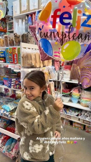 Una niña con abrigo estampado sostiene un globo de cumpleaños dentro de una tienda de cotillón con estanterías de artículos festivos