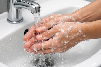 Primer plano de dos manos con espuma de jabón bajo un chorro de agua de un grifo cromado, en un lavabo blanco.