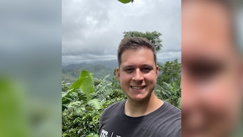 Un hombre de piel clara, cabello castaño oscuro y camiseta negra sonríe frente a un paisaje montañoso cubierto de vegetación bajo un cielo nublado