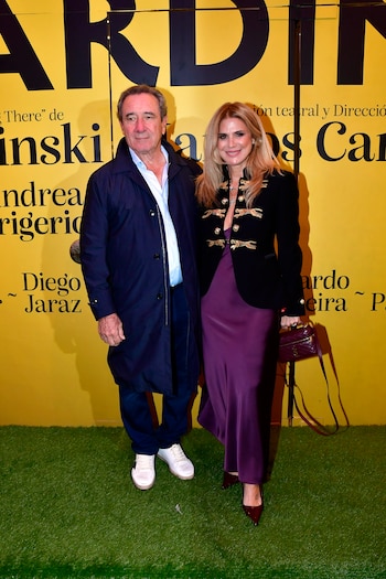 Dos personas, un hombre con gabardina azul y una mujer con blazer negro y vestido morado, posan sonrientes frente a un fondo amarillo con texto y césped artificial