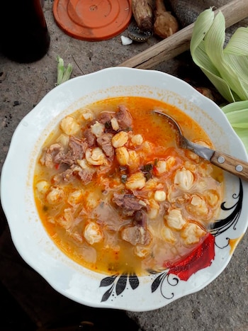 Un plato blanco decorado con locro, que contiene trozos de carne, maíz blanco y caldo anaranjado, junto a una cuchara metálica. Hay chalas de maíz al fondo