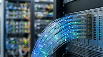 Cables de fibra óptica transparentes con destellos de luz conectados a un panel metálico junto a bastidores de servidores desenfocados.