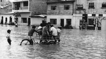 Fotografía en blanco y negro de personas sobre un triciclo empujado en una calle inundada frente a varias casas y edificios