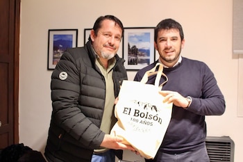 Dos hombres sostienen una bolsa de tela blanca con el texto El Bolsón 100 años frente a una pared con cuadros