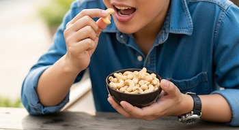 Primer plano de una persona vistiendo una camisa de mezclilla azul, llevando una castaña de cajú a la boca mientras sostiene un tazón lleno de ellas.