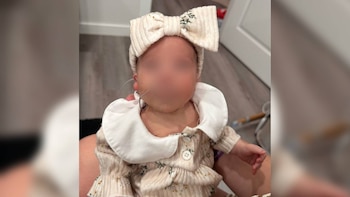 Una bebé con el rostro difuminado, vestido claro con cuello blanco, lazo en la cabeza y cánula nasal, sostenida en brazos