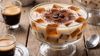 Gelatina de mosaico de café en bol de cristal, decorada con cacao en polvo y tres granos de café, acompañada de dos tazas de espresso y una cuchara sobre mesa de madera.