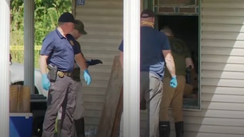 Cuatro hombres, tres con gorras y guantes azules, uno con pistola en su funda, en la entrada de una casa con número 182, un cooler y cinta amarilla