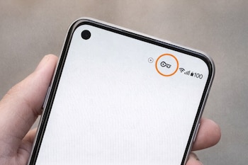 Mano sostiene un teléfono móvil con pantalla blanca, un ícono de llave en círculo naranja, indicadores de wifi y batería, y cámara frontal.