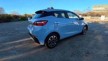 Vista tres cuartos trasera de un automóvil compacto de color celeste estacionado al aire libre en un playón despejado