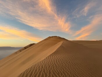 Gran duna de arena con ondulaciones detalladas, cielo azul con nubes alargadas anaranjadas y rosadas, y montañas en el horizonte