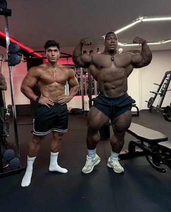 Dos hombres musculosos posan de pie en un gimnasio con máquinas de pesas, luces rojas y blancas, y un suelo negro