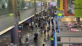 Vista aérea de un concurrido pasillo en el Aeropuerto Internacional de Miami con cientos de pasajeros caminando, letreros de puertas D27 y D1-D26, y pantallas publicitarias