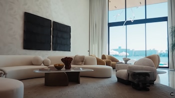 Un salón moderno y luminoso con sofás curvos crema, sillones, una mesa de centro ovalada con objetos, y grandes ventanales con vistas al mar y edificios