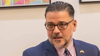 Primer plano del artista PSY con cabello oscuro peinado hacia atrás, gafas, barba, traje azul, camisa de rayas y corbata. Un mapa al fondo