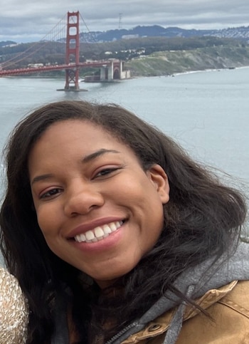 Una mujer sonriente de cabello oscuro y chaqueta ante un cuerpo de agua con el puente Golden Gate al fondo