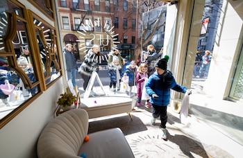 Niños y adultos buscan huevos de Pascua dentro de una tienda. Un niño con chaqueta azul camina en primer plano. El ventanal refleja edificios y la calle. Huevos de plástico en un sillón