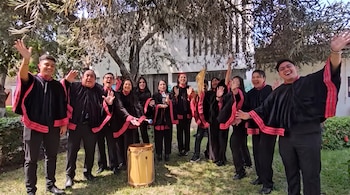 Trece personas, hombres y mujeres, sonríen y saludan con las manos levantadas. Visten capas negras con bordes rojos y rosados. Un tambor de madera en el suelo.
