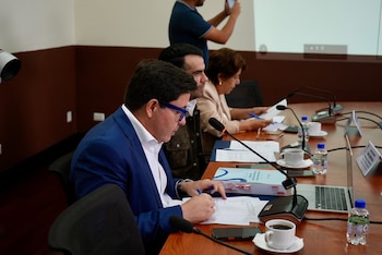 Un hombre con gafas de marco azul habla frente a un micrófono en una mesa donde hay una taza de café y una computadora portátil