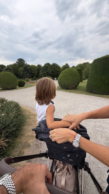 Niño sentado en cochecito, de espaldas, en un parque con senderos de grava y arbustos. Manos adultas sostienen el cochecito, una con reloj metálico, la otra con reloj y anillo