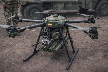 Un dron militar camuflado del Ejército de Colombia con seis rotores, una cámara y lo que parecen ser municiones, posado sobre tierra, con un soldado y un vehículo al fondo.