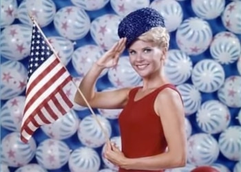 Una mujer con gorro azul y prendas rojas sostiene una bandera de Estados Unidos y realiza un saludo frente a un muro de globos