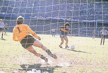 Un portero con camiseta naranja y número 1 se lanza, un jugador con camiseta azul y amarilla y número 10 patea un balón, se observa una red de fútbol, un estadio lleno de público