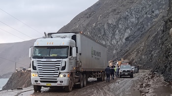 Un camión blanco transita por una vía embarrada junto a un cerro rocoso, acompañado por camionetas, personas y maquinaria pesada